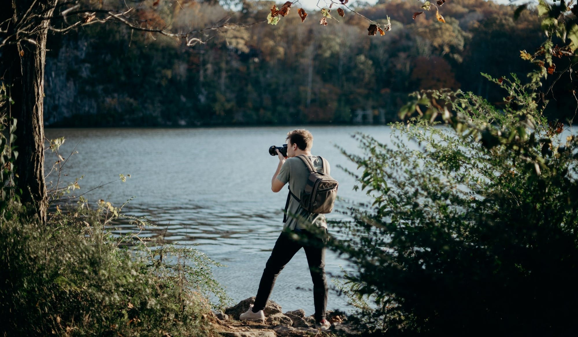 man with backpack taking picture beside lake